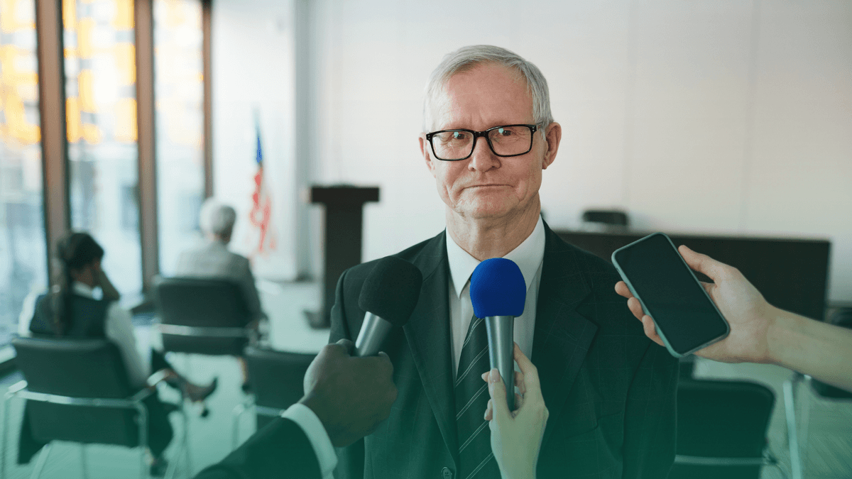 Um homem mais velho, de terno e óculos, está sendo entrevistado por repórteres segurando microfones e um smartphone em uma sala de conferências, o que evidência a importância da análise análise do teor da notícia em consulta de mídias.