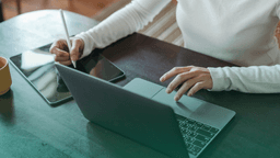 Mulher usando notebook em mesa de escritório com tablet ao lado, simbolizando a gestão de CAGED em contratações e verificação de dados profissionais.