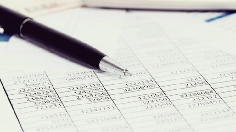 Uma caneta preta apoiada sobre uma tabela com números impressos, representando documentos financeiros ou registros de dados importantes. Ao fundo, desfocados, papéis adicionais e uma pasta podem ser vistos, reforçando a ideia de análise ou auditoria.