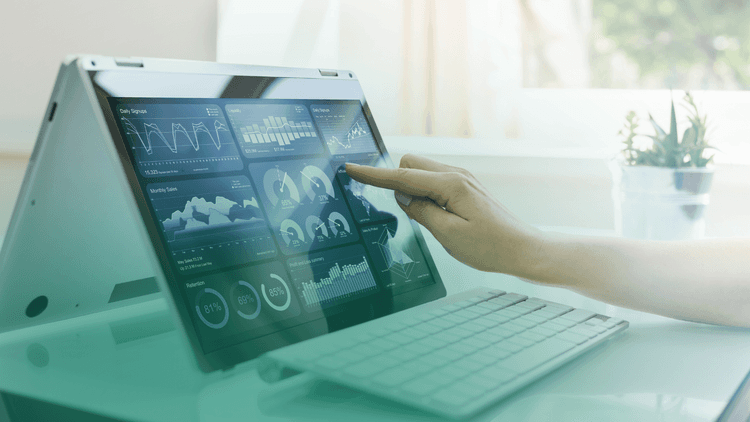 Pessoa interage com tela sensível ao toque de um notebook em formato tenda, analisando gráficos e dashboards de desempenho e indicadores operacionais em ambiente de escritório iluminado.