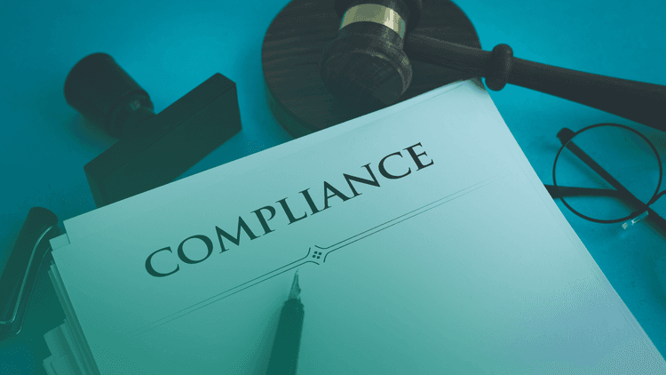 Documento com a palavra "COMPLIANCE" em destaque sobre uma mesa, ao lado de um carimbo, óculos e um martelo de juiz, representando práticas jurídicas e regulatórias.