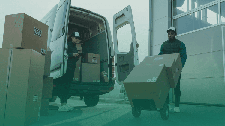 Duas pessoas, uma do sexo feminino e outra do sexo masculino carregando caixas para entrega em uma van. O homem está carregando as caixas em um carrinho de transporte.
