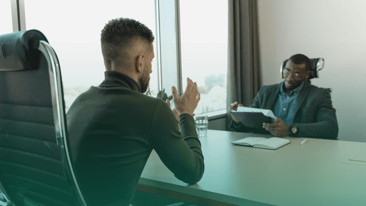 Dois homens em uma entrevista de emprego em um escritório moderno. Um deles, de costas para a câmera, está gesticulando com as mãos enquanto fala. O outro, sentado do outro lado da mesa, escuta atentamente enquanto faz anotações em uma prancheta. Há um caderno aberto e um copo de água sobre a mesa, e uma janela grande ao fundo com vista para a cidade