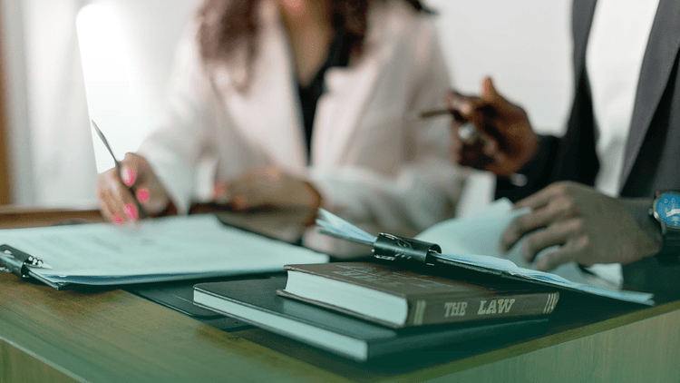 Sócios trabalhando em uma mesa repleta de documentos e livros. É um homem e uma mulher.