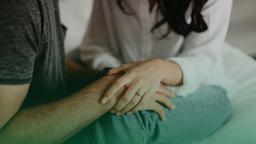 Casal sentado, a mão da mulher, usando um anel, está apoiada de forma carinhosa no braço do parceiro, transmitindo confiança e segurança.
