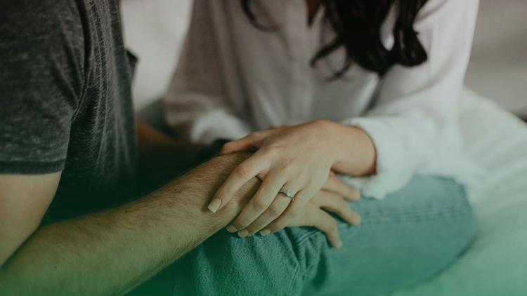 Casal sentado, a mão da mulher, usando um anel, está apoiada de forma carinhosa no braço do parceiro, transmitindo confiança e segurança.